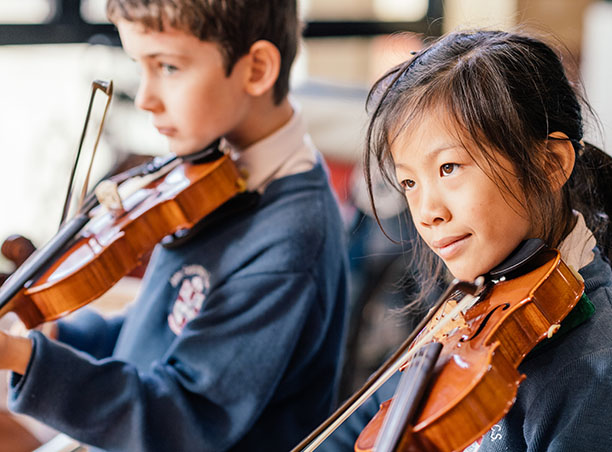 Pictured are two young students who play Music at JSRACS. Both hold a violin to their chin ready to play.