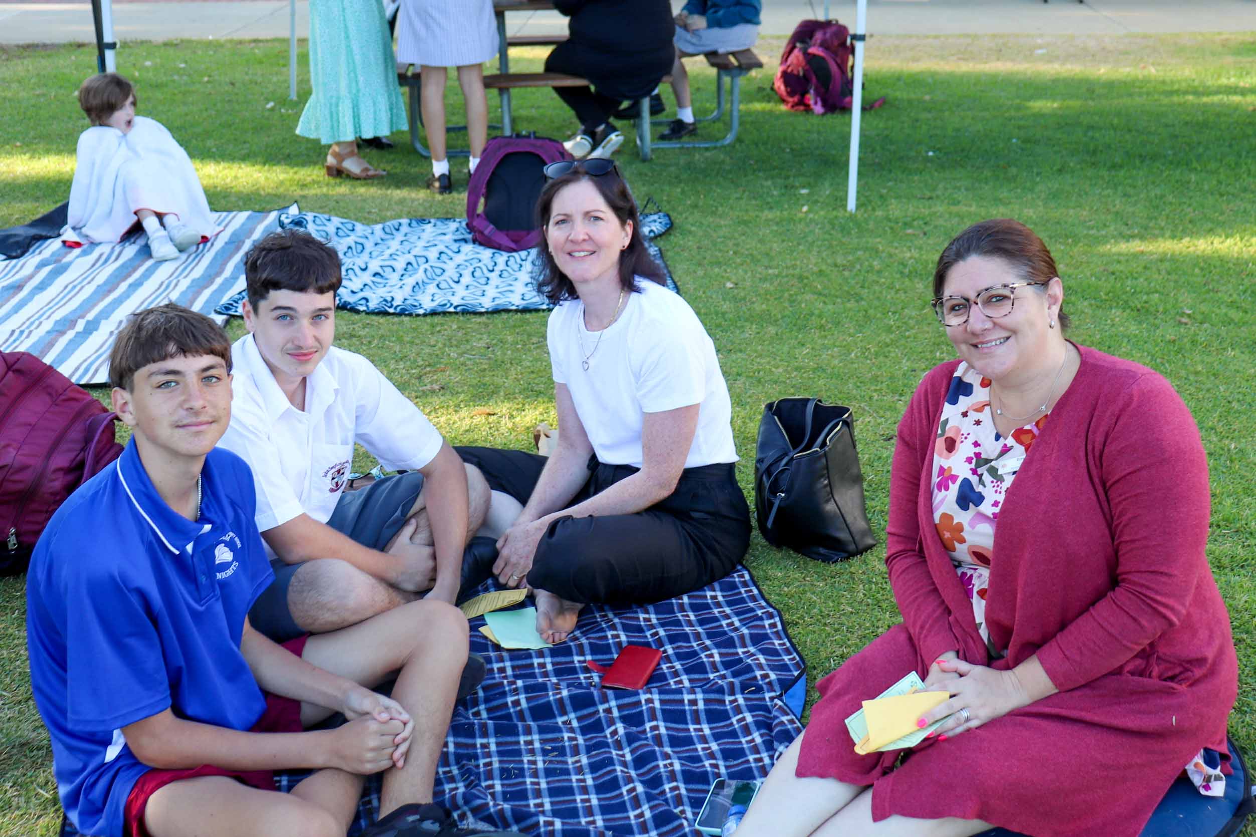 Pictured are two sons and their mothers at The Rite Journey Call and Departure Ceremony. They sit on a picnic blanket holding small sheets of paper.