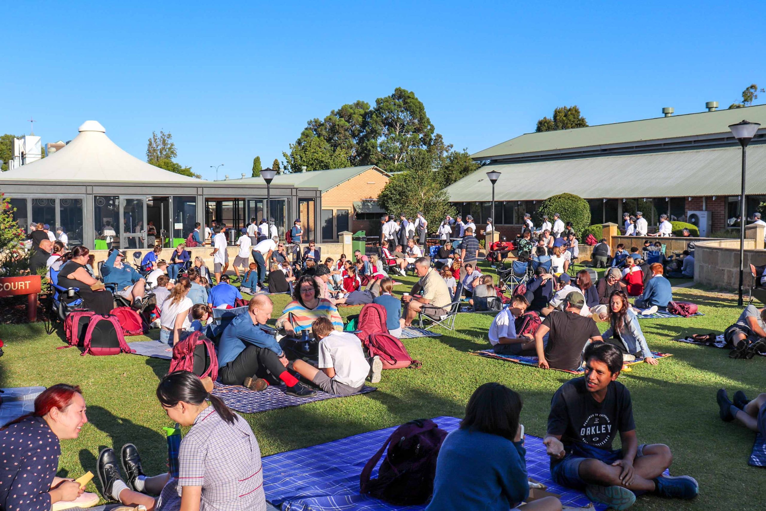 Pictured is a crowd of people attending The Rite Journey Call and Departure Ceremony. They scatter across grass on picnic mats and lawn chairs.