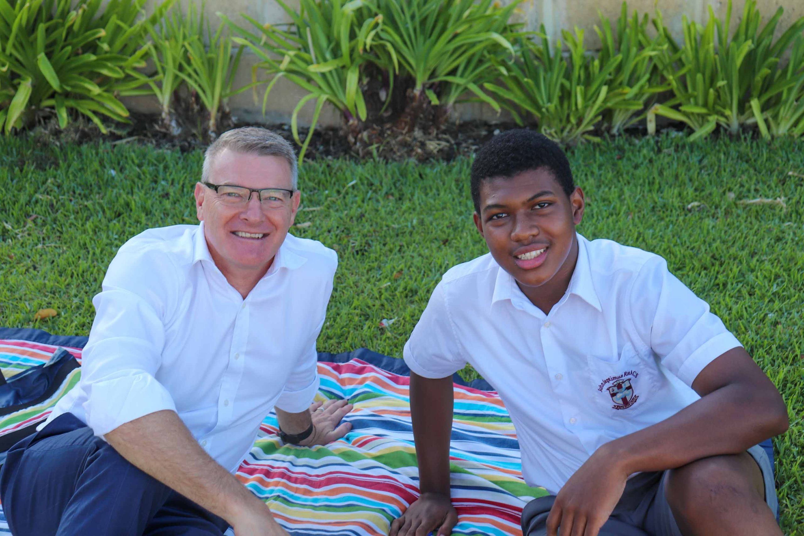 Pictured is a man and a boy at The Rite Journey Call and Departure Ceremony. They sit on a picnic rug smiling at the camera.
