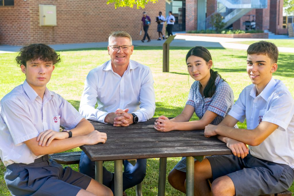 A principal and three students sit at a table facing the camera smiling. In the background you see students and teachers on their way to class.